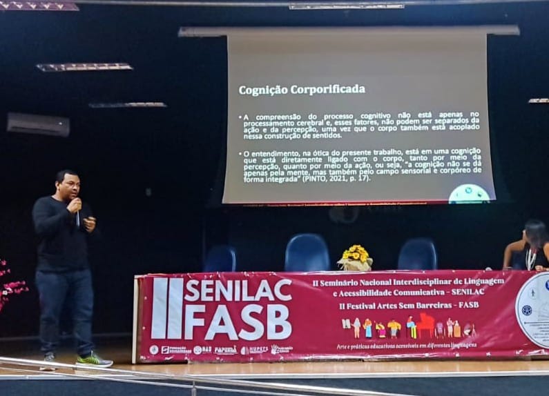 Foto no auditório do Paulo Freire. Jorge apresenta trabalho. Ao fundo o slide com sua apresentação.